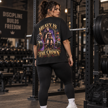Load image into Gallery viewer, A woman in a gym wearing a black graphic t-shirt with a large back print that reads 'HEAVY IS THE CROWN' and shows a powerlifting illustration. She is wearing black leggings and white sneakers.