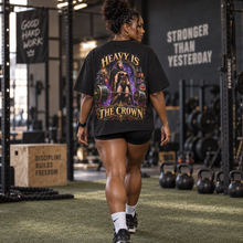 Load image into Gallery viewer, A woman from behind in a gym wearing a black graphic t-shirt with a large illustration and the text 'HEAVY IS THE CROWN'.