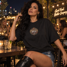 Load image into Gallery viewer, A woman seated at an outdoor restaurant table at night wearing a black graphic T-shirt with a decorative chest print.
