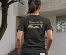 Load image into Gallery viewer, A woman with a pony tail, standing with her hands in her pockets, wearing a "Transformation Never Diminishes Beauty" Black T-Shirt