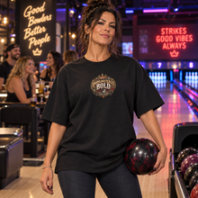 Load image into Gallery viewer, A woman in a bowling alley wearing an oversized black graphic t-shirt with a gold and white 'UNAPOLOGETICALLY BOLD' crown logo. She is holding a black and red marbled bowling ball.
