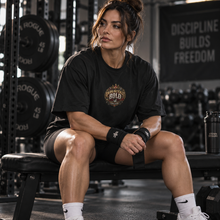Load image into Gallery viewer, A woman sits on a weight bench in a gym wearing a black t-shirt with a gold and white 'UNAPOLOGETICALLY BOLD graphic on the chest, black athletic shorts, and white socks. The background shows weight racks and a sign that reads 'DISCIPLINE BUILDS FREEDOM'.
