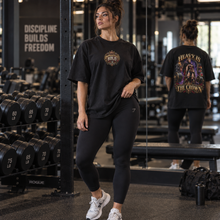 Load image into Gallery viewer, A woman in a gym wearing a black oversized t-shirt with a gold 'Crown' graphic on the front and a large back graphic that reads 'HEAVY IS THE CROWN', paired with black leggings and white sneakers. She is standing in front of a mirror showing the back of the shirt, with dumbbells and gym equipment in the background.

