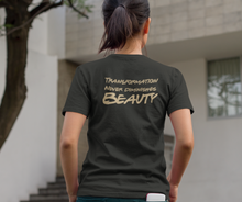 Load image into Gallery viewer, A woman with a pony tail, standing with her hands in her pockets, wearing a &quot;Transformation Never Diminishes Beauty&quot; Black T-Shirt 

